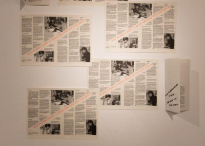 Seven leaflets displayed on a white wall, five are flat, two are folded. The leaflets feature black and white photographic images and text with some fluorescent orange text.