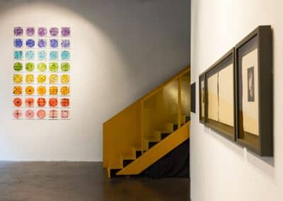 Installation view of exhibition, 40 square prints hung in a grid fashion on a white wall in the background beside a staircase painted yellow. Prints in black frames are hung on a wall in the right foreground.