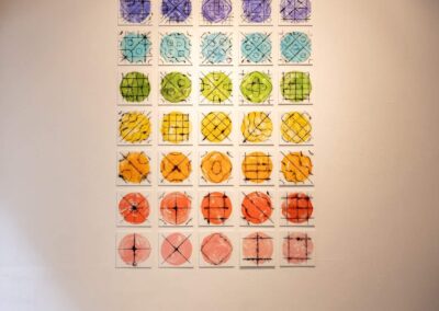 Forty square prints hung on a white wall in a grid format. The prints feature violet, indigo, blue, green, yellow, orange, red, and pink circles with black lines and shapes on top.