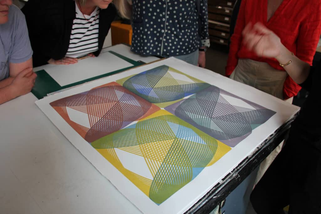 Image with coloured print on a table., surrounded by 4 figures. Only the bodies of the humans are visible.